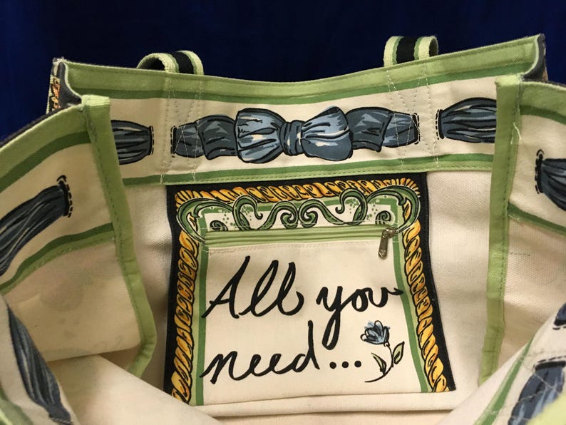 May include: A cream-coloured tote bag with light green trim and a blue bow pattern along the top edge. The interior features a central zippered pocket with a yellow rope-style border and the black printed text "All you need..." with a small blue flower.