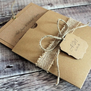 May include: A rustic wedding invitation with a brown paper envelope, a burlap ribbon, and a twine bow. The tag attached to the bow reads "A&M 12.05.18".