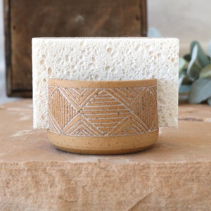 May include: A tan ceramic sponge holder with a geometric design and a white sponge. The holder has a cylindrical shape with a textured surface and a pattern of white lines. The sponge is rectangular and fits in the holder.