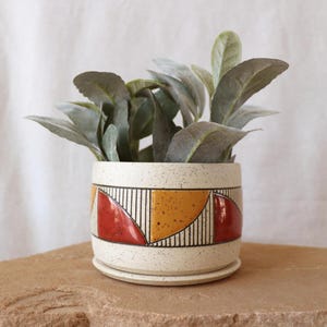 May include: A speckled ceramic planter with a geometric design in red, yellow, and black. The planter holds a leafy green plant. The planter sits on a matching saucer, and the background is a neutral color.