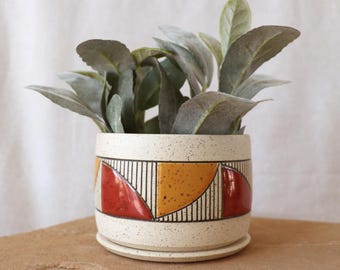 Könnte beinhalten: Ein gesprenkelter Keramik-Pflanztopf mit einem geometrischen Design in Rot, Gelb und Schwarz. Der Topf enthält eine grüne, belaubte Pflanze. Der Topf steht auf einer passenden Untertasse. Der Hintergrund ist neutral.