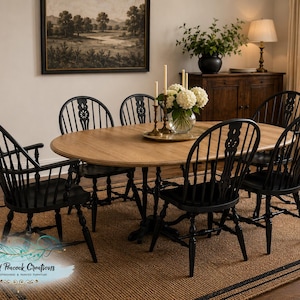 May include: An oval wooden dining table with a natural light brown finish and a pedestal base, surrounded by six black spindle-back chairs. The table is set with a glass vase containing white hydrangea flowers and two tall, thin taper candles.