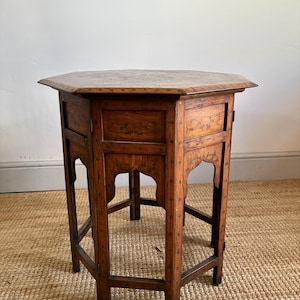 May include: An octagonal wooden side table featuring a dark brown finish with intricate inlaid patterns. The table has six legs connected by lower stretchers and arched cutouts on each side panel, showcasing detailed floral motifs and geometric accents.