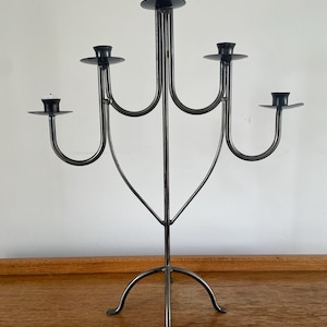 May include: A black metal candelabra with five candle holders. The candelabra has a central stem and a curved base with three feet. The candle holders are circular and are arranged symmetrically.