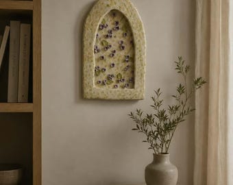 Può includere: Una placca da parete ad arco con una cornice testurizzata di colore chiaro, caratterizzata da un centro incassato riempito con piccoli decori floreali viola e foglie verdi. L'oggetto è esposto su una parete sopra una superficie in legno con libri e un vaso.