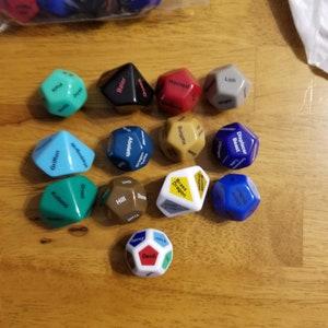 May include: A set of 12 colorful dice with different fantasy creature names printed on each side. The dice are arranged in a circle on a wooden surface. The dice are made of plastic and are about 1 inch in diameter.