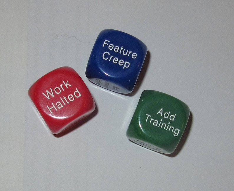 May include: Three dice with different colored backgrounds and white text. The dice are red, blue, and green. The text on the dice reads "Work Halted", "Feature Creep", and "Add Training".
