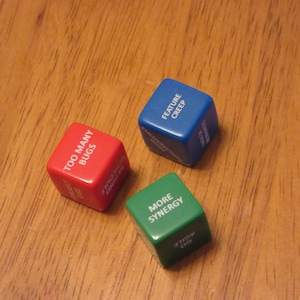 May include: Three dice with different colored backgrounds and white text. The red die says "Too Many Bugs", the blue die says "Feature Creep", and the green die says "More Synergy".