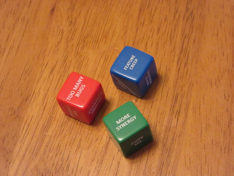 May include: Three dice with different colored backgrounds and white text. The red die says "Too Many Bugs", the blue die says "Feature Creep", and the green die says "More Synergy".