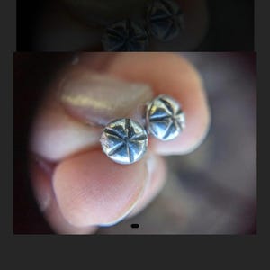 May include: A pair of small, silver-colored stud earrings. Each earring has a rounded shape with a starburst design etched into the surface. The earrings are held between fingers, with a blurred background.