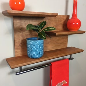 May include: A wooden wall shelf with three shelves and a towel bar. The shelf has a plant in a blue and white patterned pot, an orange glass vase, and an orange glass bowl. A red towel with a cheetah print hangs on the towel bar.