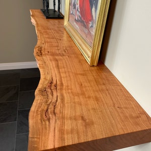 May include: A live edge wooden shelf with a warm reddish-brown tone and visible natural wood grain patterns. The shelf is mounted to a wall and holds a gold-framed painting depicting a figure in a red garment and a black wire sculpture.