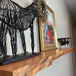 May include: A wooden shelf with a live edge displays decorative items. A black wire lion sculpture with gold accents sits next to a framed painting and a small ceramic bowl with a colorful design. The shelf is made of light brown wood.