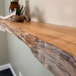 May include: A natural wood shelf with a live edge, featuring a unique grain pattern and knots. The shelf is mounted on a wall and has a light brown finish.