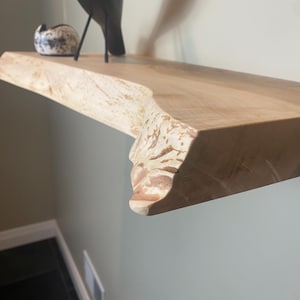 May include: A natural wood floating shelf with a live edge. The shelf is light brown with a lighter, textured edge. A black metal bird sculpture and a small speckled ceramic piece are displayed on the shelf.