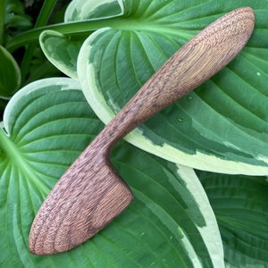 Może przedstawiać: Drewniany nóż do masła w ciemnobrązowym kolorze z zakrzywionym uchwytem i lekko spiczastym ostrzem. Nóż leży na zielonym liściu z białymi brzegami.