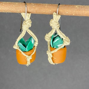 May include: A pair of handmade earrings featuring small terracotta pots with green succulent plants, hanging from macrame string.