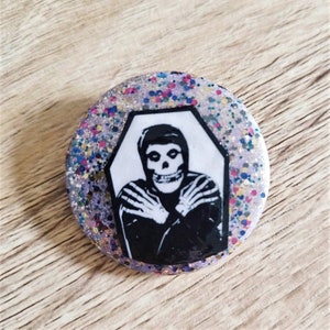 以下が含まれることがあります： キラキラと輝く紫色の背景に、棺の中に骸骨の顔をした人がいる白黒の画像が描かれた丸いボタン。画像は白い八角形に囲まれています。