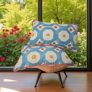 May include: Two decorative cushions with a geometric pattern in blue, white, yellow, and orange. The cushions have a coral-coloured trim and are placed on a chair with black legs. The background shows a lush green garden with red flowers.