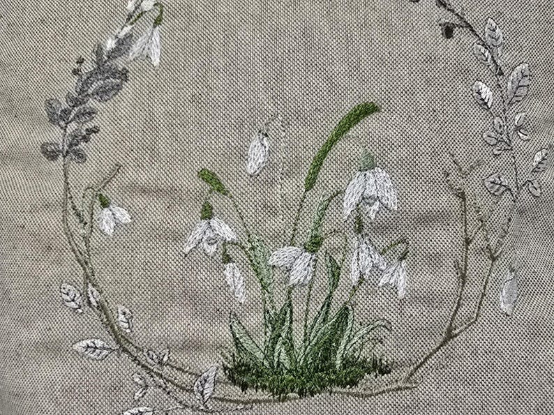 May include: Embroidered design of a wreath of white snowdrops with green leaves on a beige linen fabric.