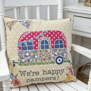 Peut inclure: Un coussin carré beige à pois blancs orné d'une applique représentant une caravane rouge à motifs floraux avec des guirlandes, de l'herbe brodée en vert et l'inscription We're happy campers ! en lettres gris foncé sur le bas.