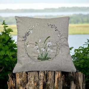 May include: A gray linen pillow with an embroidered design of white snowdrops and green leaves in a circular wreath.