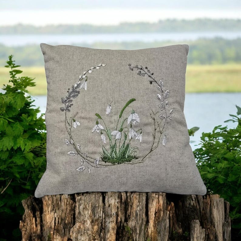 May include: A gray linen pillow with an embroidered design of white snowdrops and green leaves in a circular wreath.
