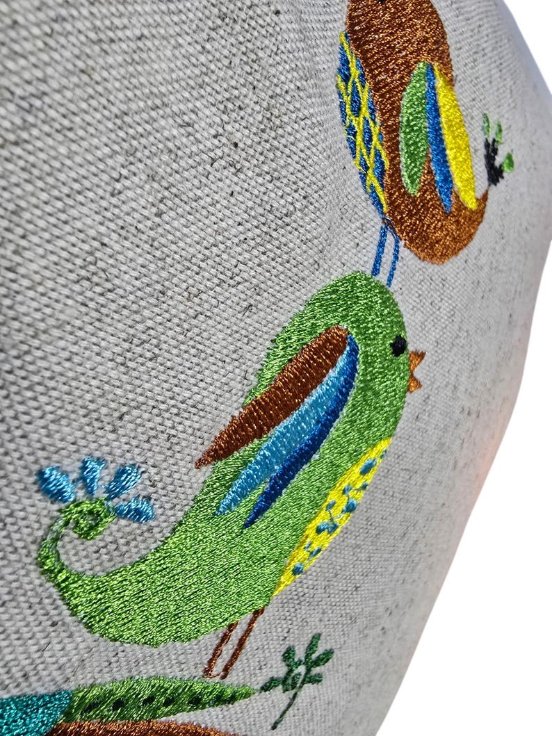 May include: Embroidered birds on a light gray fabric background. The birds are green, blue, yellow, and brown. The birds are perched on a branch with green leaves.