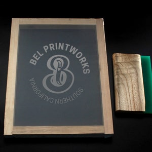 May include: A wooden screen printing frame with a gray screen and a white logo that says "BEL PRINTWORKS SOUTHERN CALIFORNIA" with a stylized number 8 and a knife inside the number. A wooden squeegee with a green rubber blade is next to the frame.
