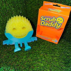 May include: A yellow Scrub Daddy sponge with a smiling face, blue arms and legs, and an orange box with the Scrub Daddy logo. The box reads "Scratch Free" and "Resists Odors". The sponge is designed for cleaning.