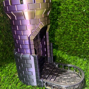 May include: A 3D-printed dice tower in shades of purple, black, and gold, resembling a castle tower. The tower features a brick pattern, a small balcony, and a detailed design. The tower is set against a green background.