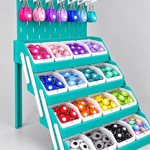 May include: A teal tiered retail display stand featuring a pegboard top with hanging personal safety alarms and four rows of white bins filled with various coloured personal safety alarms in shades of pink, purple, blue, green, orange, yellow, black, and white.