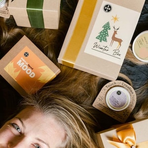 Könnte beinhalten: Eine Frau mit blonden Haaren ist von Geschenkboxen umgeben, die in braunes Papier mit grünem, goldenem und gelbem Band verpackt sind. Die Boxen haben Etiketten mit dem Text "A Zen A", "Winter Box", "Dobro je #good MOOD" und "A Touch of Croatian Nature".