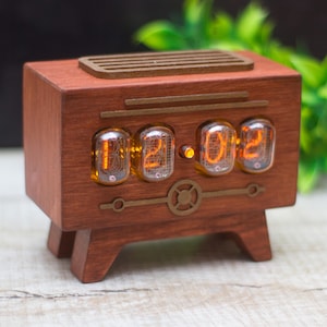 May include: A wooden digital clock in a retro radio design. The clock features four glowing orange digits displayed in glass tubes. The clock is made of dark brown wood and has a decorative speaker grill on top.
