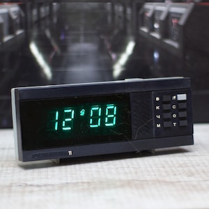 May include: A black digital alarm clock showing the time 12:08 in green digits. The clock has a rectangular design with buttons on the right. A small number 11 is visible at the bottom left.