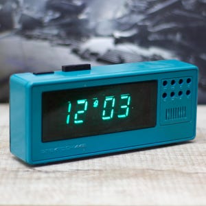 May include: A teal digital alarm clock displaying 12:03 in green numerals. The clock has a rectangular shape with a speaker and button details on the side. The text "ЭЛЕКТРОНИКЕ" is printed below the time display.