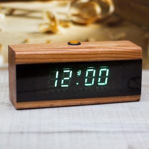 May include: A rectangular digital alarm clock with a natural wood grain finish and a black front display panel. The screen shows the time 12:00 in bright green LED digits, accompanied by a small bell icon. A single orange button sits on the top.