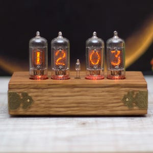 May include: A wooden digital clock with four glowing orange Nixie tubes displaying the time. The clock has a rectangular wooden base with decorative metal corner accents. The tubes are encased in clear glass.
