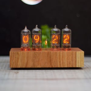 May include: A nixie tube clock featuring four glass vacuum tubes displaying the orange glowing numbers 6, 9, 2, and 2, mounted on a rectangular light-colored wooden base with a small neon indicator bulb positioned between the second and third tubes.