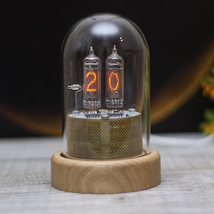 May include: A nixie tube clock featuring two glowing orange vacuum tubes displaying the number 20, encased within a clear glass dome. The tubes sit on a circular base with a textured olive green fabric wrap and a light-colored wooden bottom.