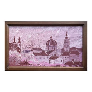 May include: An oil painting of a cityscape in shades of pink and white, framed in a brown wooden frame. The artwork depicts buildings with domes, spires, and rooftops, with textured brushstrokes creating a sense of depth and movement.
