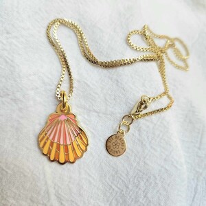 May include: A gold chain necklace with a seashell pendant. The seashell is pink, orange, and yellow with a white interior. The pendant is attached to a small gold charm that says "See Beachy Designs".