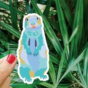 Könnte beinhalten: Ein Aufkleber mit einem blauen, grünen und gelben Präriehund, der auf seinen Hinterbeinen steht. Der Präriehund hat einen weißen Umriss und ist vor einem grünen, belaubten Hintergrund platziert.