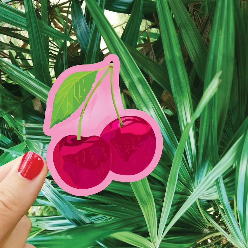 K&ouml;nnte beinhalten: Ein Aufkleber mit zwei Kirschen mit einem gr&uuml;nen Blatt und Stielen, umrandet in Rosa. Die Kirschen sind tiefrot. Der Hintergrund ist &uuml;ppiges gr&uuml;nes Laub.