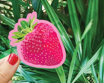 Puede incluir: Una pegatina de fresa rosa brillante con hojas verdes y un contorno rosa. La pegatina está colocada sobre un fondo de exuberante follaje verde. La imagen también muestra una mano con esmalte de uñas rojo.