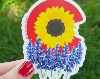 May include: A sticker featuring a red, yellow, and blue design. The design is a stylized outline of the state of Colorado with a sunflower in the center and bluebonnets at the bottom. The sticker has the text "@beebeacheydesigns" on the side.