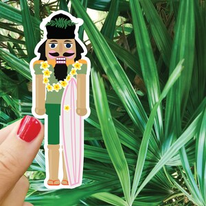 Pode incluir: Uma figura de quebra-nozes com um colar de flores e segurando uma prancha de surf rosa. A figura usa uma camisa verde, shorts marrons e uma barba preta. O fundo é uma planta verde frondosa.