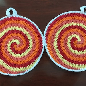 May include: Two crocheted pot holders with a spiral pattern in shades of orange, red, and yellow. The pot holders have a white border.