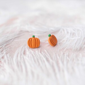 May include: A pair of orange pumpkin-shaped earrings with green stems. The earrings are on a white fluffy surface.