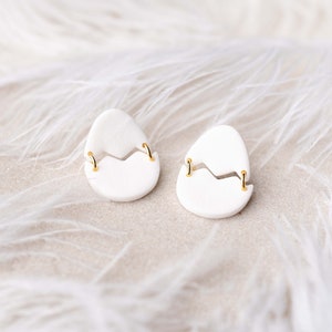 May include: A pair of white ceramic earrings shaped like cracked eggs with gold accents.
